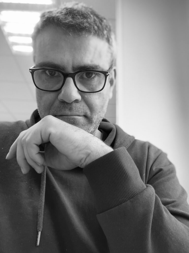 Portrait en noir et blanc de Laurent Ménéla portant des lunettes, en sweat à capuche, la main posée devant le visage dans une pose réfléchie.